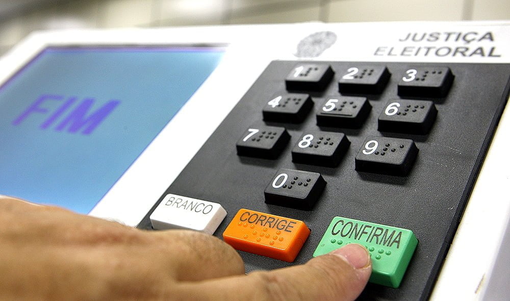 Foto mostra dedo apontado para a tecla confirma da urna eletrônica. No visor do urna aparece escrito a palavra "Fim"