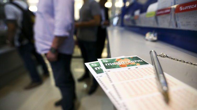 Canhoto de apostas com caneta em cima. Ao fundo, fila para fazer apostas.
