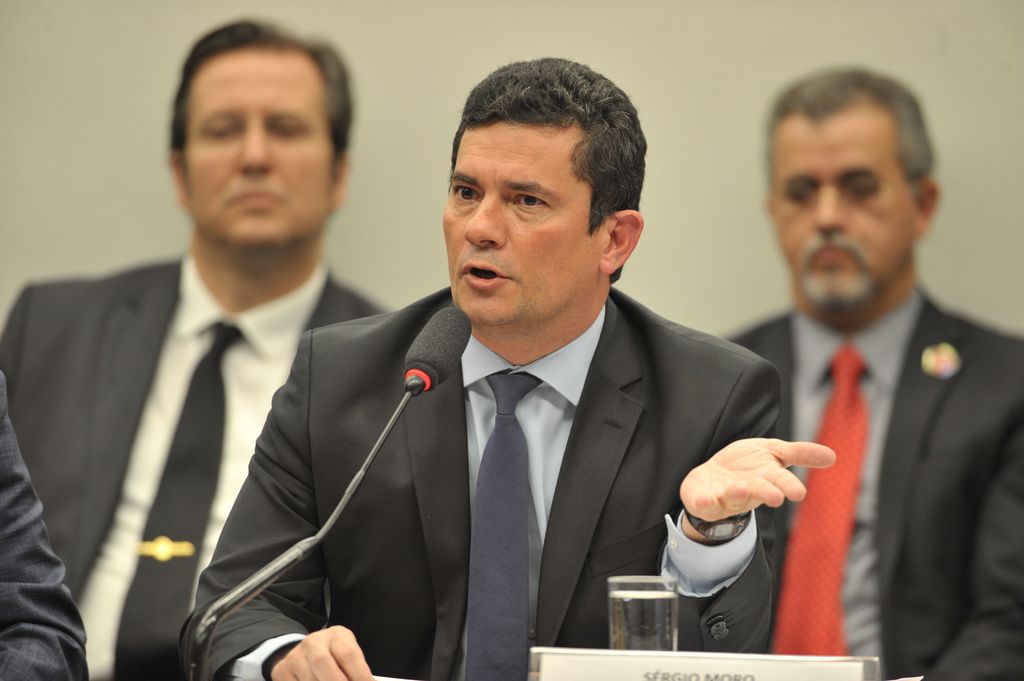 Moro faz gesto com a mão esquerda enquanto fala ao microfone. Ao fundo, dois homens acompanham fala.