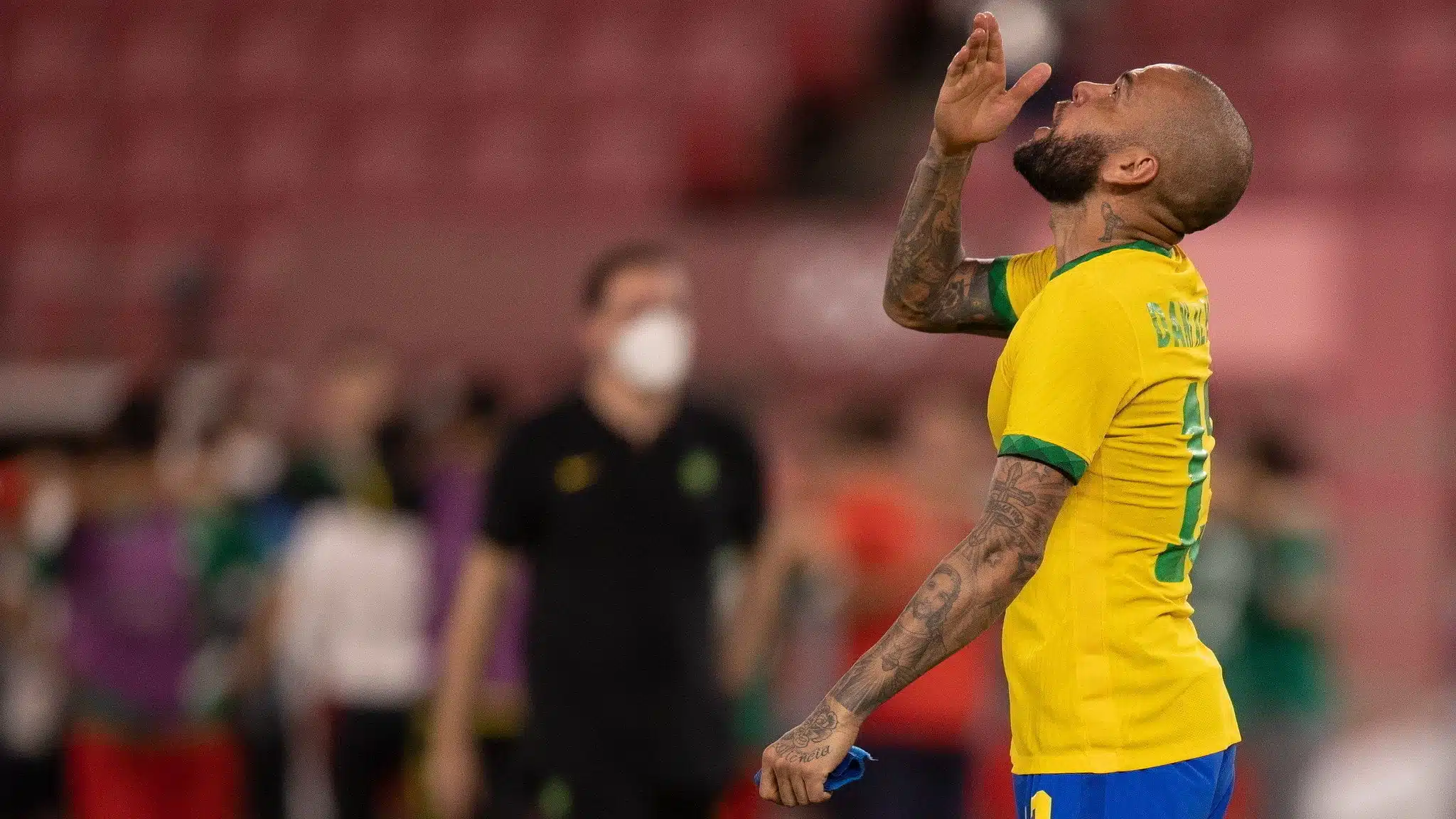 Daniel Alves com a camisa da seleção brasileira, bebe água enquanto ao fundo aparecem outras pessoas desfocadas pela camera.