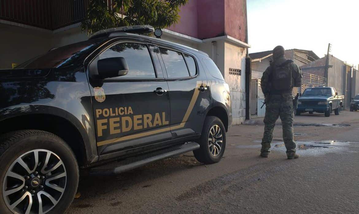 Viatura da Polícia Federal estacionada e policial armado ao lado.