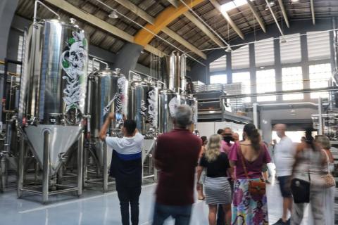 Cilindros de cerveja ao fundo. Pessoas, sem resolução de imagem, observam a estrutura dos cilindros