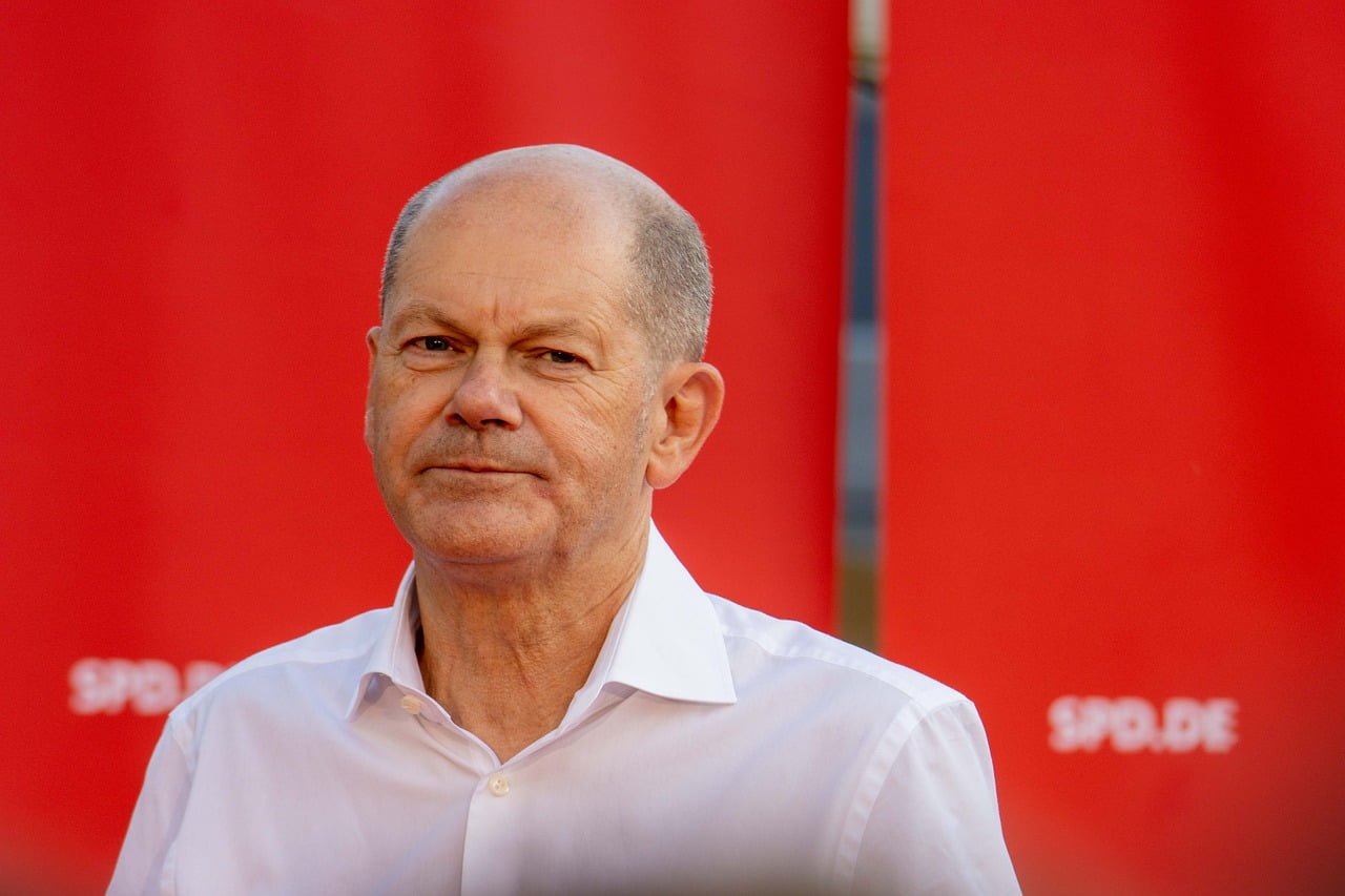 Olaf Scholz de camisa social branca, com fundo vermelho.
