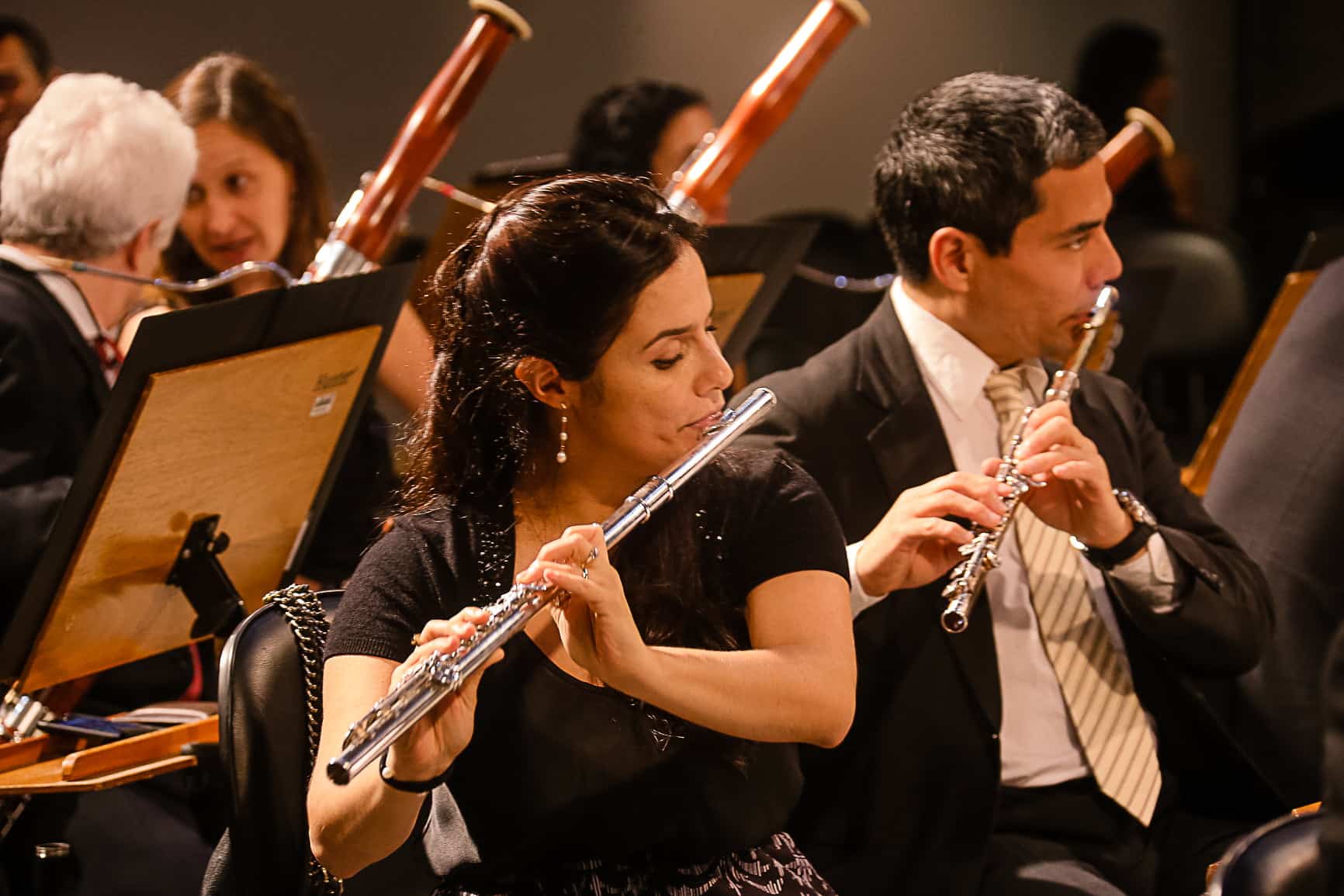 Músicos da Orquestra da USP tocam flauta. No primeiro plano está uma mulher branca, cabelos pretos lisos presos, com a flauta na boca. No segundo plano, um homem branco, cabelos grisalhos, de terno preto e gravata listrada também com a flauta na boca tocando.