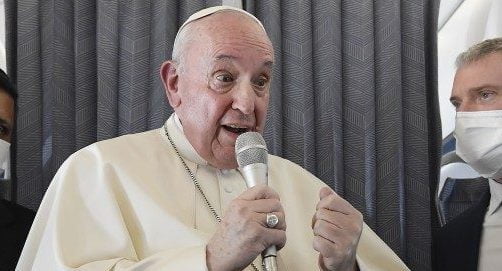 Papa Francisco segurando o microfone. Enquanto fala, é observado por dois assessores que usam máscara e terno e gravata.