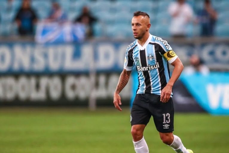 Jogador Rafinha com o uniforme do Grêmio no gramado. Ao fundo, poucos torcedores na arquibancada.