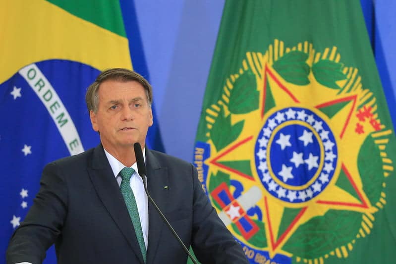 Jair Bolsonaro, homem de pele branca, cabelos grisalhos na lateral da cabeça e cabelos penteados para o lado. Ao fundo bandeiras.