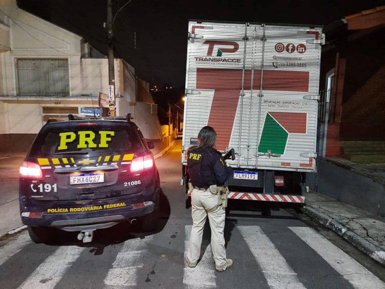 Viatura da Polícia Rodoviária Federal ao lado do caminhão. Agente da PRF, com arma longa, aparece olhando o caminhão, sobre a faixa de pedestres.