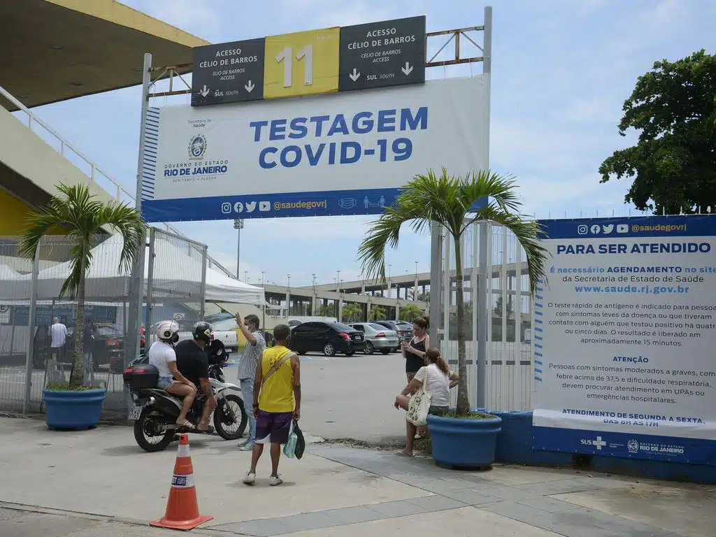 Entrada do estacionamento de local para testagem de covid. Pessoas na porta paradas e faixa com a frase "testagem covid-19" no alto.