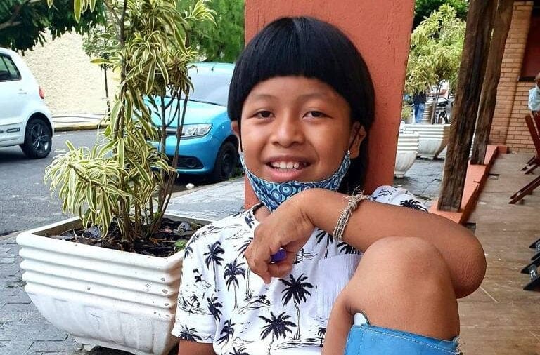 Davi Xavante, menino de cabelos lisos, olho levemente puxado, sentado e sorrindo.