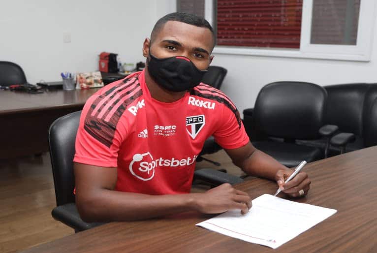 Jogador Nikão, usando uniforme do São Paulo Futebol Clube, segura a caneta diante de uma folha de papel colocada sobre a mesa. O jogador está com máscara.