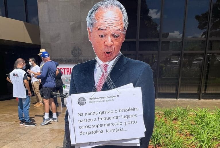 Imagem de Paulo Guedes em papel olhando para baixo e com um cartaz preso ao pescoço. Ao fundo, servidores durante protesto.