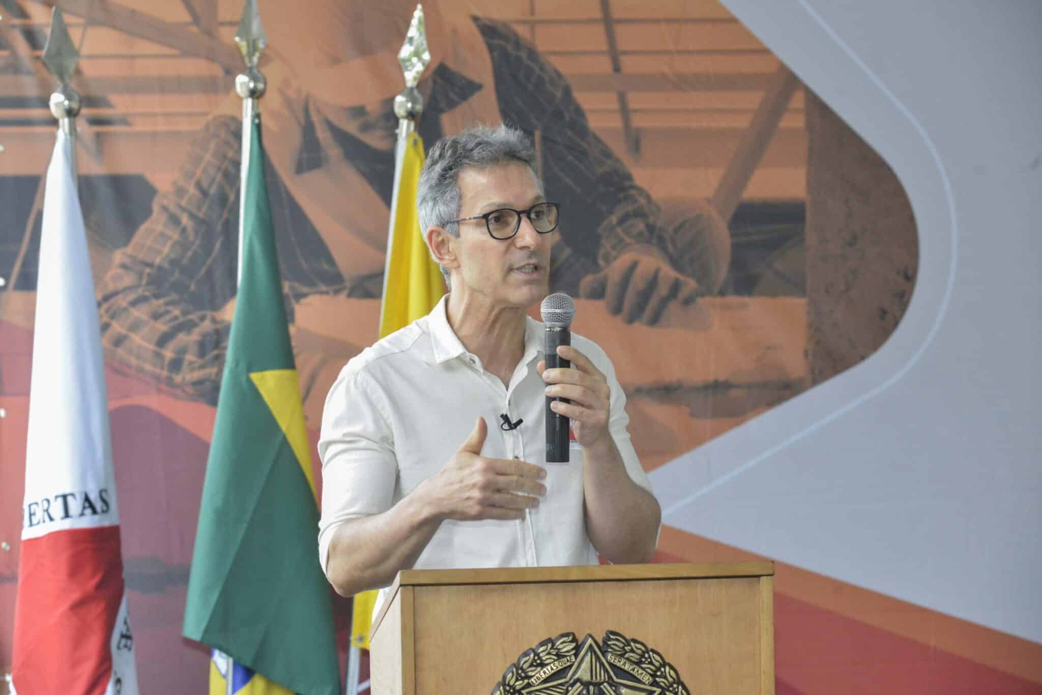 Romeu Zema, homem de pele branca, cabelo grisalho, segura microfone enquanto discursa. Ao fundo, bandeiras, inclusive do Brasil.