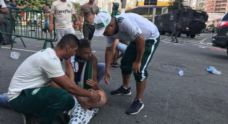 Torcedor caído no chão e outros torcedores ao redor, abaixados, conversando com a vítima enquanto espera pelo resgate.