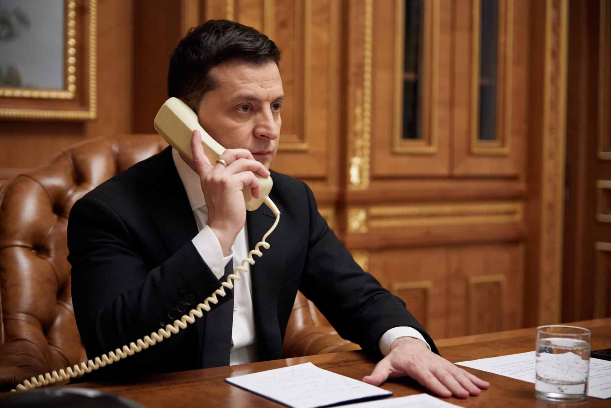 Volodimyr Zelensky, presidente da Ucrânia, fala ao telefone sentado à mesa. A mão esquerda está estendida sobre a mesa, onde há folhas de papeis.
