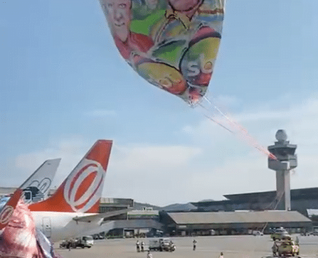 Balão colorido com imagens de personagens do SBT se aproxima do chão enquanto outra parte já está sobre a calda de um avião estacionado.