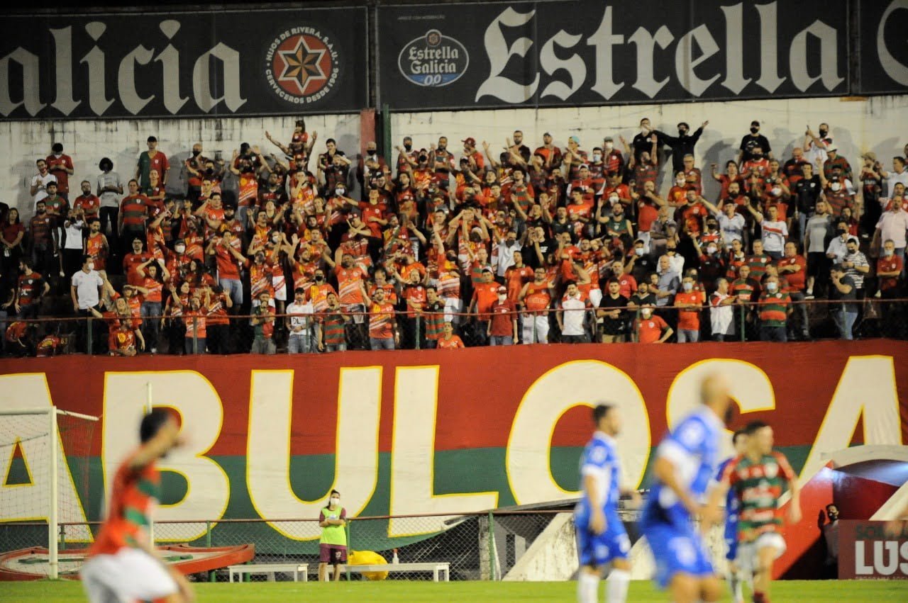 Torcida faz festa na arquibancada enquanto jogadores, no primeiro plano da imagem olham para o lado direito da foto, dentro do campoi.