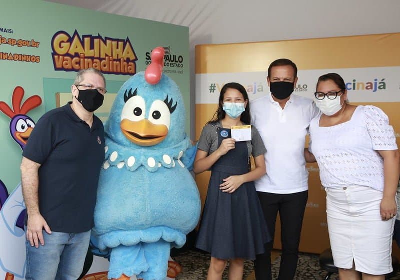 Secretario de saúde do estado, jean gorinchteyn, posa para foto ao lado de personagem infantil, uma ave do desenho Galinha pintadinha. Também estão na foto uma criança vacinada, o governador João doria e uma mulher, vestida de branco.