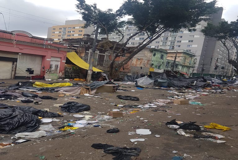 Imagem mostra local onde ficava a cracolândia, com roupas no chão espalhadas pela rua e calçada, imóveis próximos. Ao fundo, novos condomínios de prédios construídos na região para habitação popular.