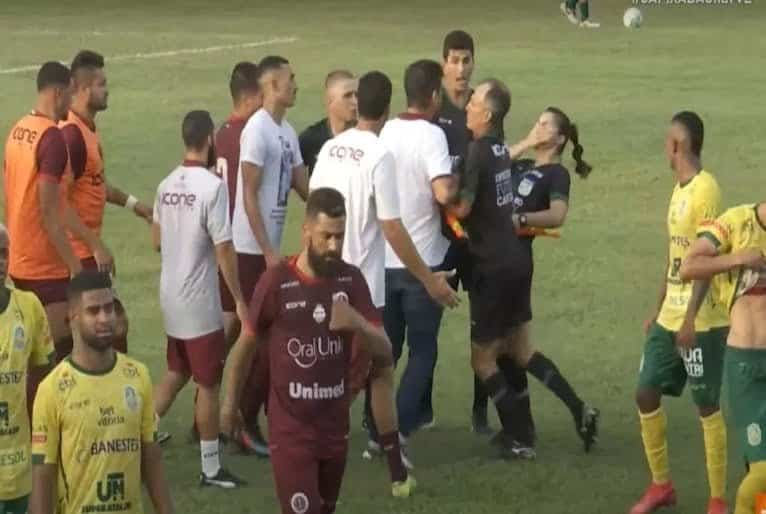 Imagem mostra bandeirinha com a mao no rosto logo após ser agredida. O técnico aparece em meio a jogadores aglomerados próximo ao árbitro.