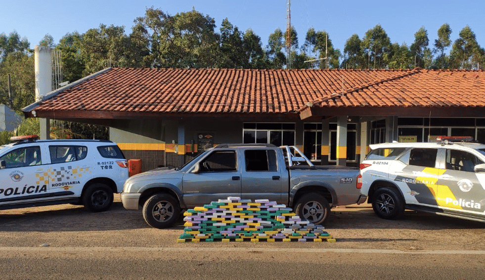 Droga empilhada ao lado de caminhonete. Nos cantos direito e esquerdo, aparecem duas viaturas da Polícia Rodoviária, responsável pela apreensão.
