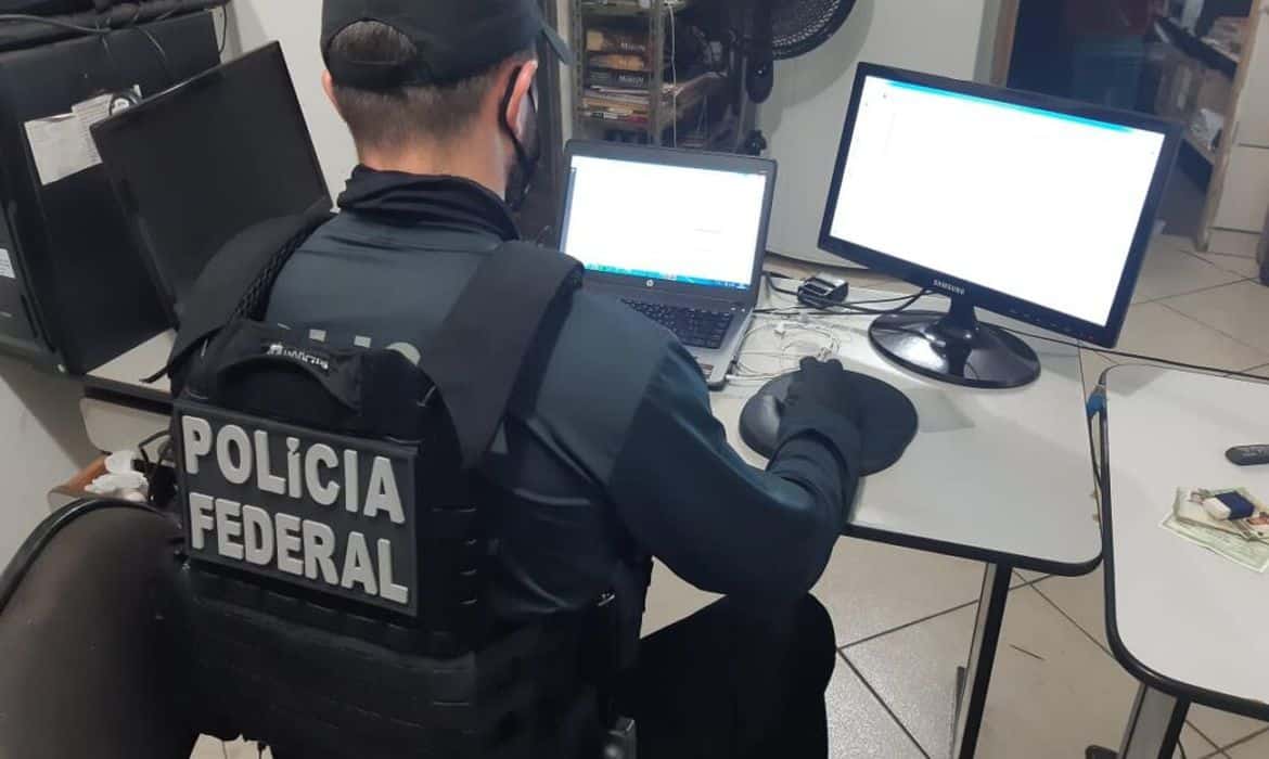 Agente da polícia federal, sentado diante de computador. Nas costas dele, a farda traz o nome "Polícia Federal".