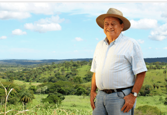 Alysson Paolinelli, com chapéu, diante de terras com árvores ao fundo.