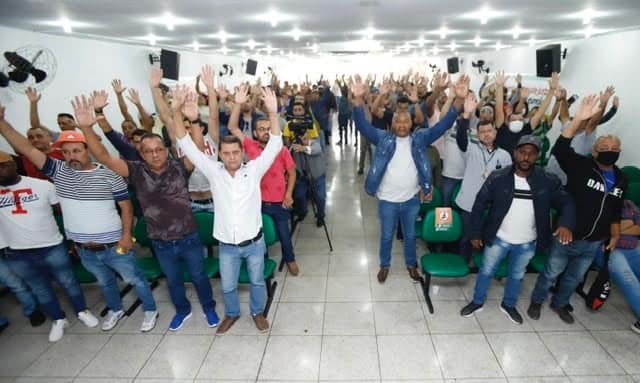 Motoristas e cobradores com os braços para o alto durante votação sobre manifestação. Todos estão dentro de um auditório.