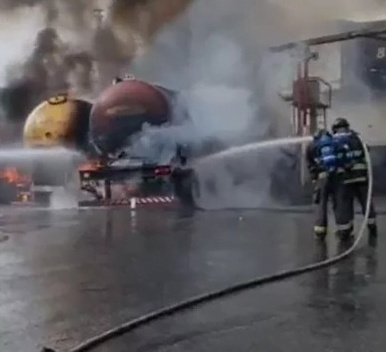 Bombeiros lançam água sobre caminhões. É possível ver chamas na parte de baixo dos caminhões e muita fumaça.