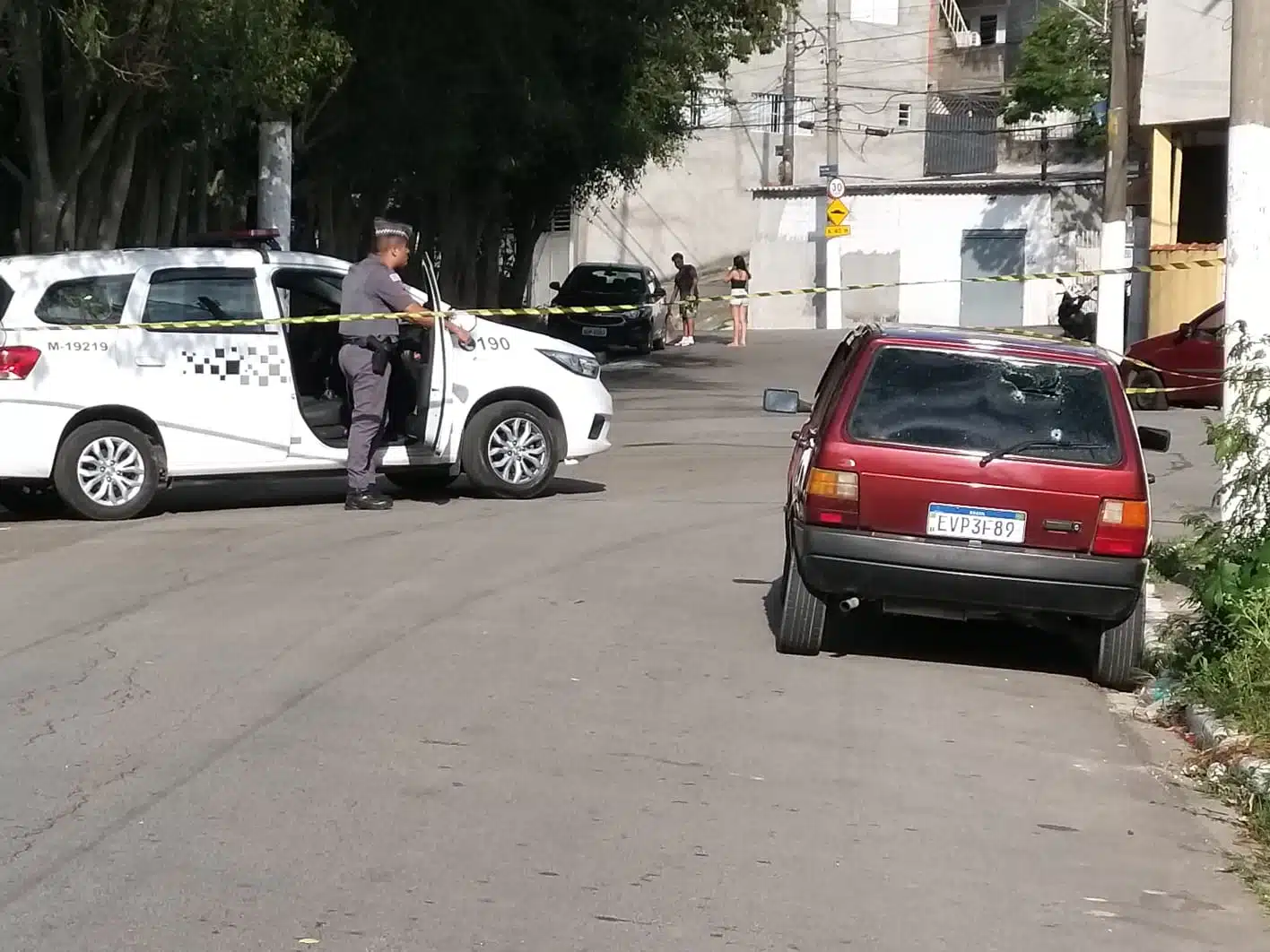 Carro estacionado, isolado com fita de emergência usada pela polícia, com marcas de tiros no vidro traseiro. Mais à frente, do outro lado da rua, está uma viatura da polícia militar e um soldado da pm com a porta do passageiro aberta e uma das pernas dentro da viatura.