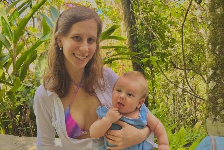 Jéssica Mayara Ballock sorri para foto com o filho no colo. Bebê aparece com a mãozinha na boca.