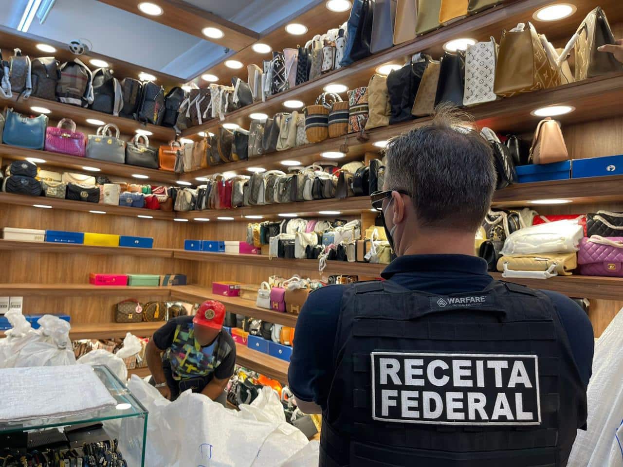 Agente da REceita Federal diante de prateleira chega de acessórios. Mulher aparece manipulando objetos diante dele.