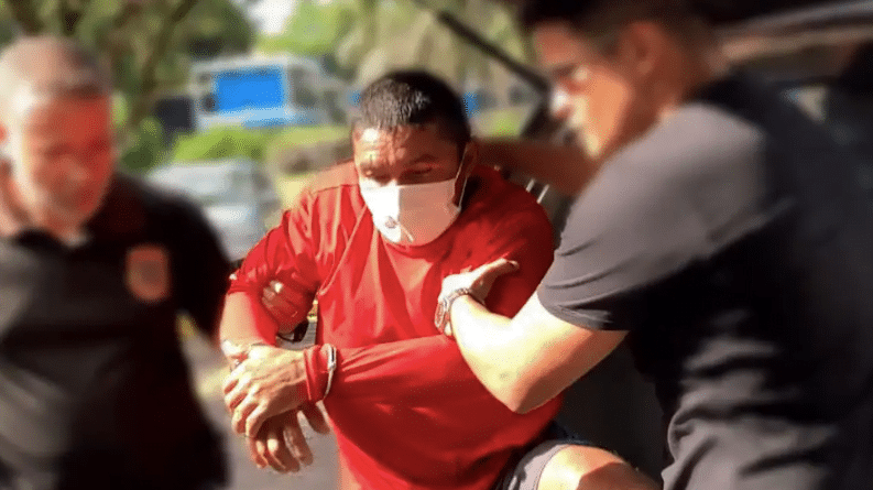 Oseney sendo transferido. Ele é retirado da parte de trás da viatura por dois agentes. Está algemado e usando máscara.