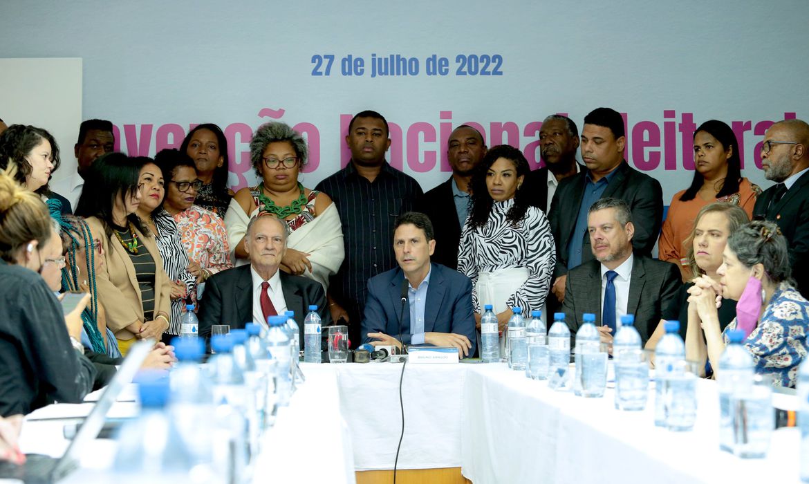 Integrantes dos partidos, sentados à mesa, enquanto outros permanecem atrás, em pé.