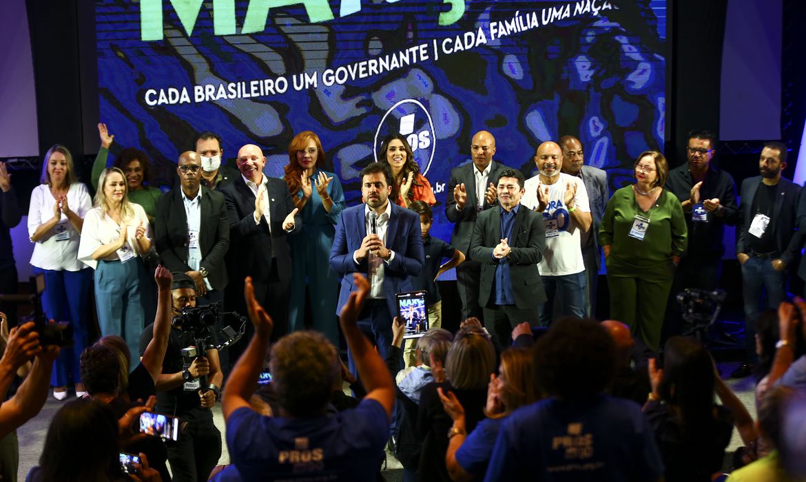 Foto mostra convenção com várias pessoas no palco, enquanto outras assistem a apresentação.