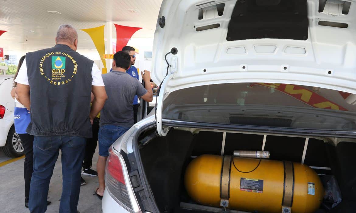 Carro aparece com porta-malas aberto, com equipamento para gás gnv dentro. Ao lado do carro, um fiscal da ANP conversa com pessoas.