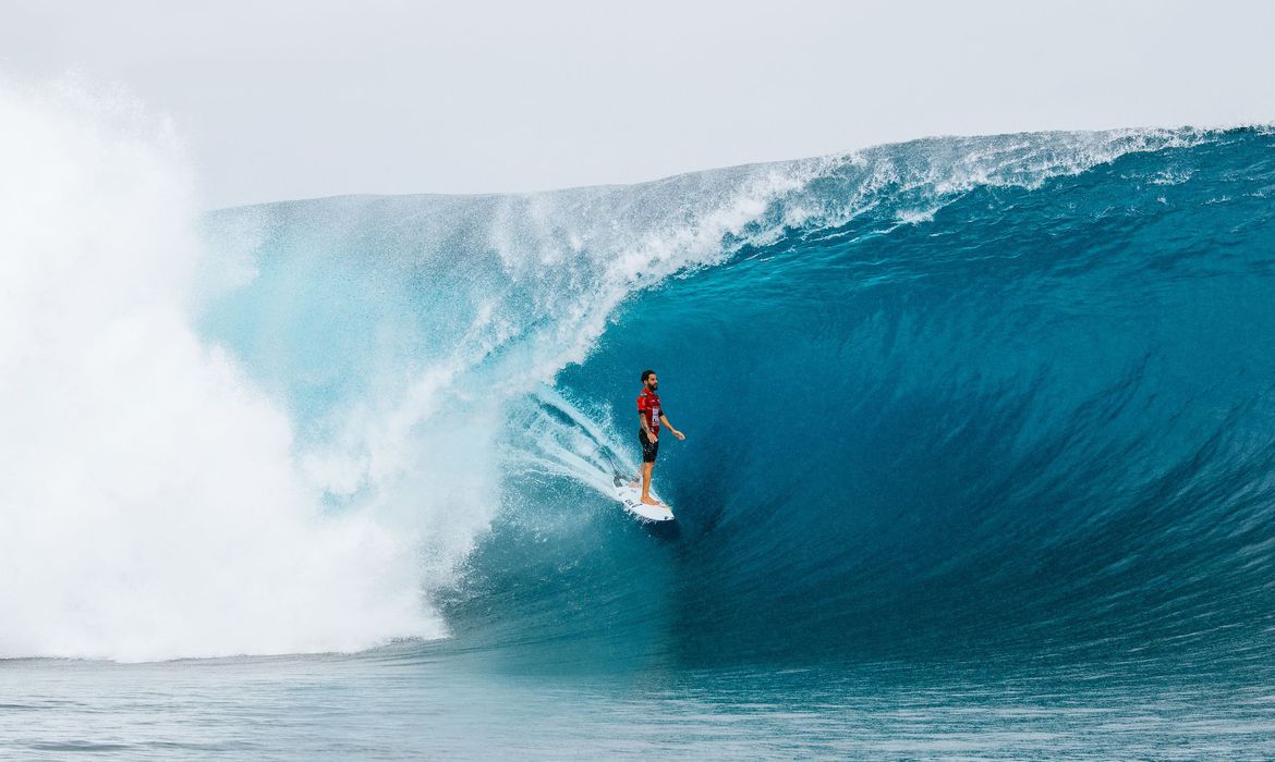 Italo Ferreira aparece em pé sobre prancha de surf dentro de uma onda. Ele desliza sobre a água enquanto a onda passa por cima dele, criando um funil.