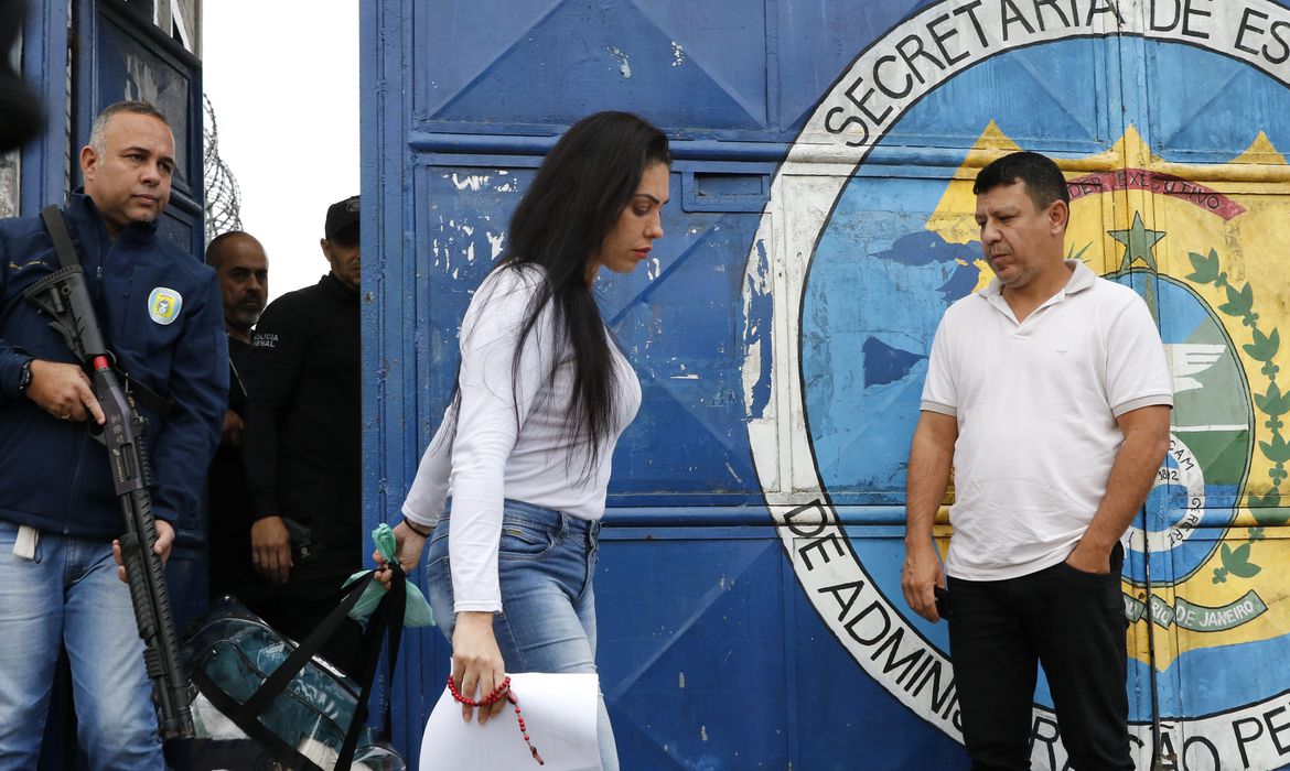 Monique deixa a prisão de cabeça baixa. Ela segura na mão uma folha de papel. Ao fundo, um agente penitenciário aparece armado e um homem, com a mao no bolso, está parado ao lado do portao.