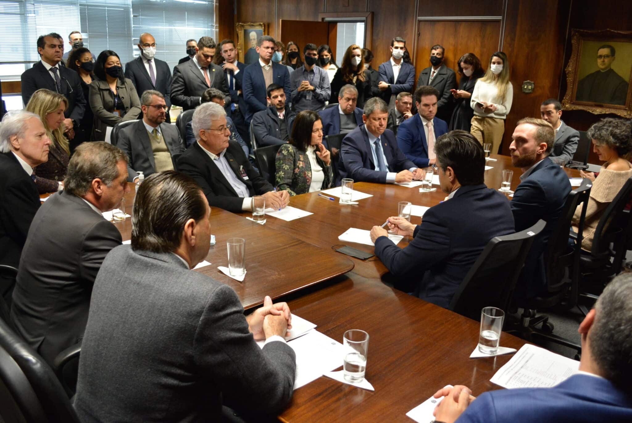 Candidados e assessores sentados, à mesa, enquanto conversam. Outros assessores aparecem ao fundo em pé.