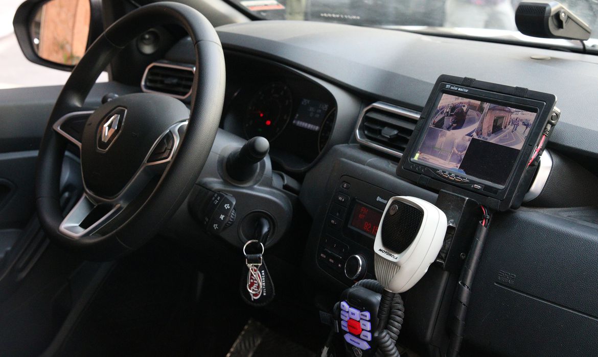 Parte interna de carro tem pequeno visor com imagem de três câmeras projetadas. Aparecem ainda o volante, a chave no contato e o rádio comunicador da viatura.