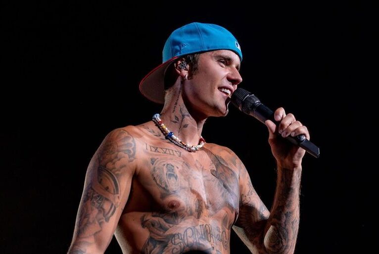 Justin Bieber. Rapaz de pele clara e coberta por tatuagens segura o microfone enquanto canta com boné azul na cabeça virado para trás.