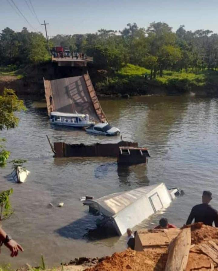 Foto mostra caminhões e carros dentro d'água e estrutura da ponte destruída após desabamento. Homem, na margem, observa os veículos.
