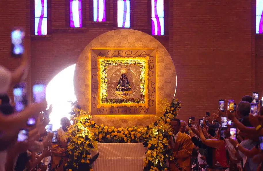 Andor com imagem de Nossa Senhora Aparecida entra na igreja. Fieis apontam celular dos dois lados do corredor.