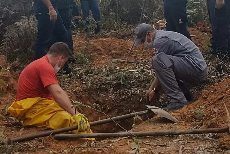 Bombeiros escavam local onde cemitério clandestino foi encontrado. Foto mostra dois bombeiros, um sentado à margem da vala e outro abaixado. Aparece ao lado também uma pá.