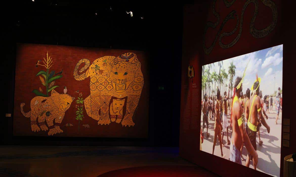 Foto mostra parte da exposição, com parede coberta por ilustração e um telão com imagens de índios.