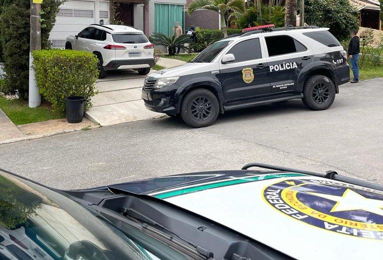 Carros da Polícia Civil e da Receita parados em frente a casa de alto padrão. Um carro de luxo está estacionado na porta da casa.