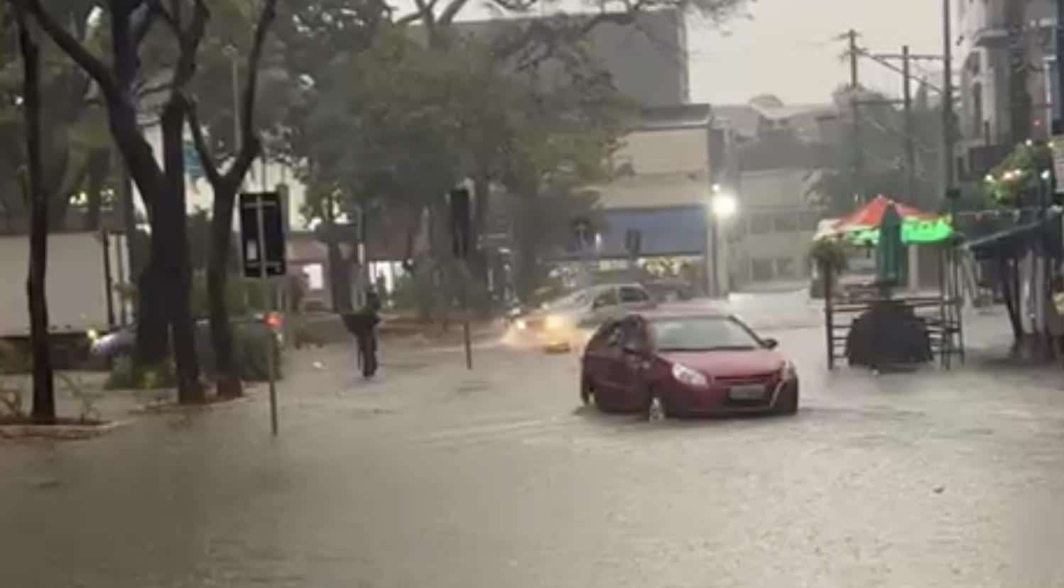 Carro dá marcha ré em rua alagada após chuva. Asfalto desaparece debaixo d'água. Ao fundo, carro passa por ponto alagado.