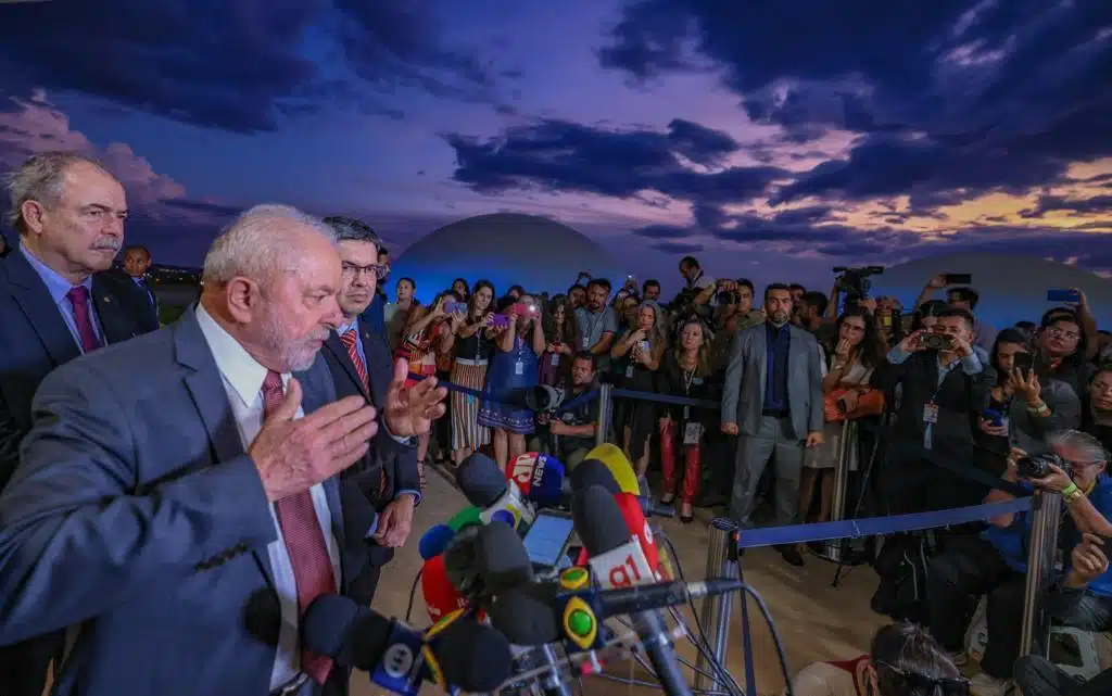 Lula fala diante de vários microfones. Ao fundo, público acompanha entrevista. Ao lado dele, outros apoiadores do PT e partidos que integram a Frente Ampla.