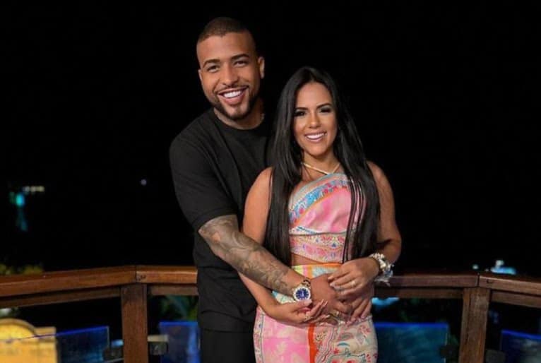 Rodrigo, home de pele escura, tatuagem cobrindo o antebraço direito, abraça por trás Hynara, mulher de cabelos lisos, longos.