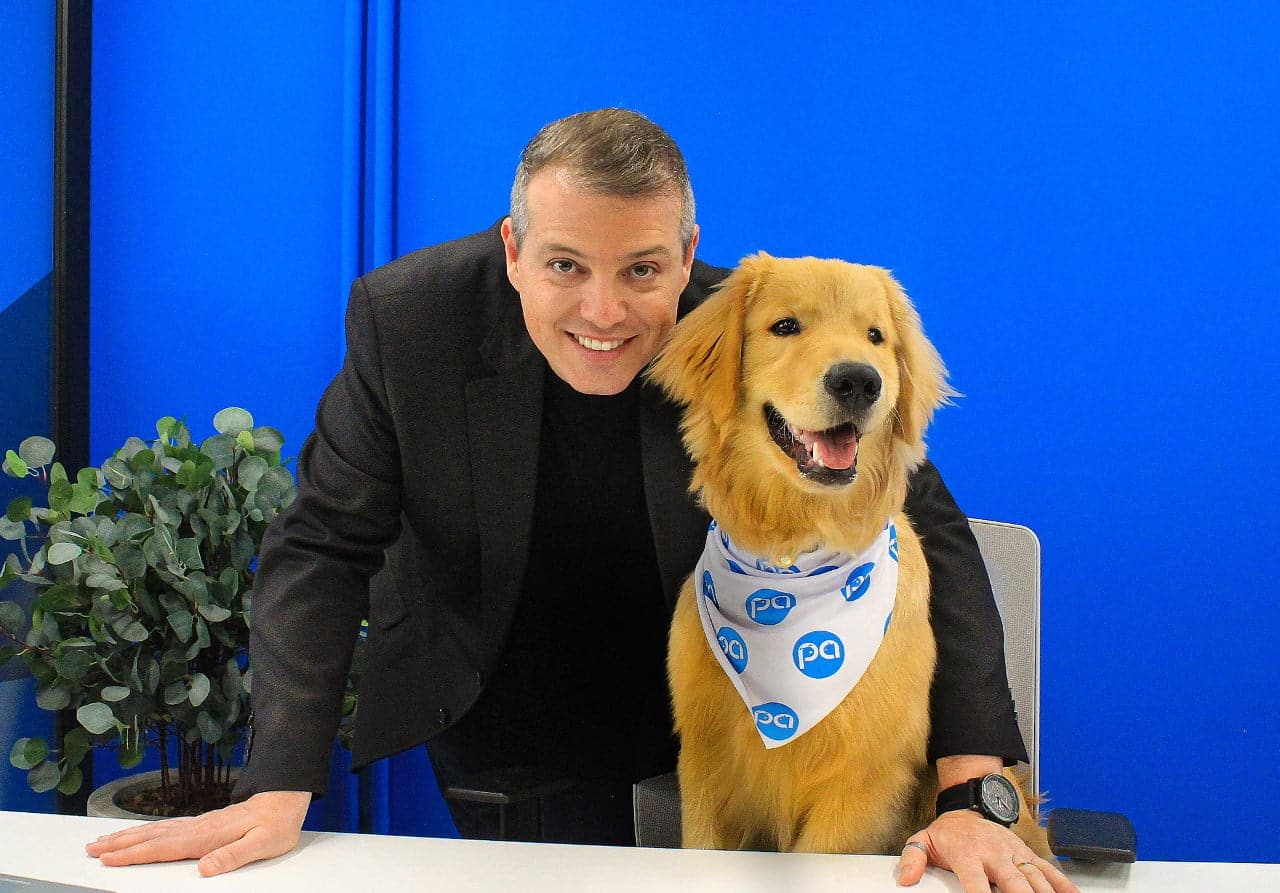 Pedro Svacina, homem de pele clara, cabelos grisalhos, penteados para o lado, se apoia com as duas mãos sobre uma mesa para ficar com o rosto próximo de um cachorro. O animal usa lenço no pescoço e está encostado na mesa na altura do executivo.